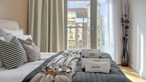 Une chambre d’hôtel avec un lit, des oreillers, un plateau avec une tasse et des fruits, et une fenêtre donnant sur des immeubles.