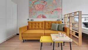 Een kamer met een mosterdkleurige bank, een kleine gele tafel met twee kopjes, een houten bed en een kleurrijk schilderij met een maritiem thema aan de muur.
