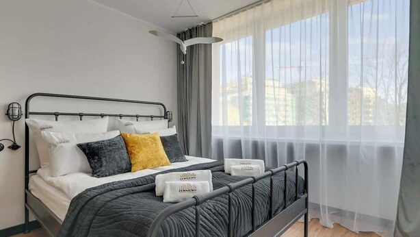Een moderne slaapkamer met een metalen bedframe, een grijs gewatteerde spreidek en uitzicht op gebouwen door dunne gordijnen.