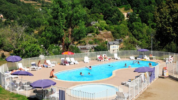 Una piscina con sillas de descanso y sombrillas, rodeada por una cerca blanca, con colinas y árboles verdes al fondo.