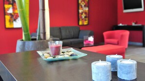 Una habitación con una pared roja, un sofá, una silla roja, una mesa con una bebida y velas, y un televisor en la pared.