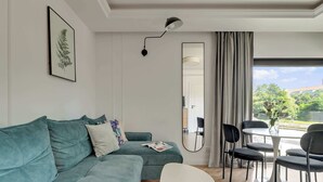 Una sala moderna con un sofá color turquesa, una mesa de centro blanca, una mesa de comedor redonda con sillas, un espejo grande y vistas a zonas verdes.
