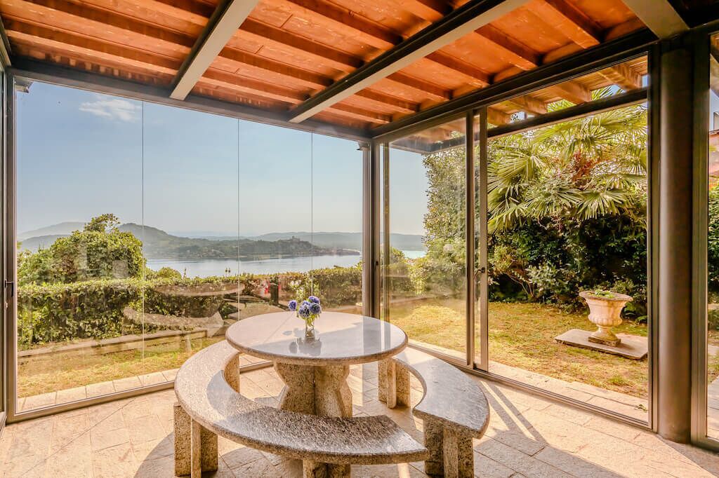 Um alpendre coberto com uma mesa e banco de pedra, amplas portas de vidro e vista para um lago e colinas.