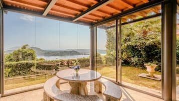 Um alpendre coberto com uma mesa e banco de pedra, amplas portas de vidro e vista para um lago e colinas.