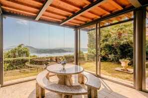 Um alpendre coberto com uma mesa e banco de pedra, amplas portas de vidro e vista para um lago e colinas.