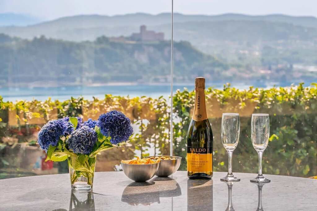 Uma mesa com uma garrafa de vinho VALDO, dois copos, uma tigela de massa e um vaso de flores, com uma vista panorâmica ao fundo.