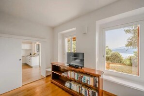 Uma sala de estar moderna com uma unidade de entretenimento de madeira, televisão e amplas janelas com vista para um jardim e montanhas.