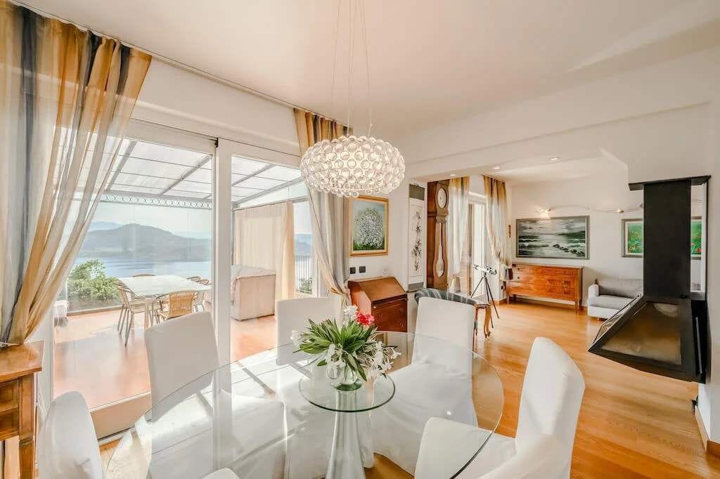 Una sala de estar moderna con una mesa de comedor de cristal, sillas blancas, una lámpara de araña y vistas al exterior.