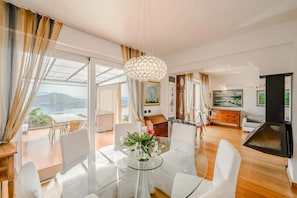 Una sala de estar moderna con una mesa de comedor de cristal, sillas blancas, una lámpara de araña y vistas al exterior.