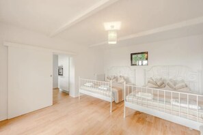 Quarto com duas camas de metal branco, uma porta branca e chão de madeira.