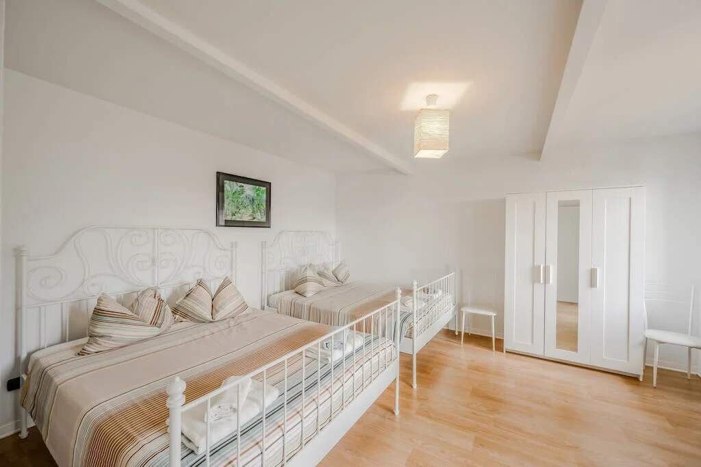 Quarto com duas camas, um guarda-roupa branco e uma pequena mesa de cabeceira.