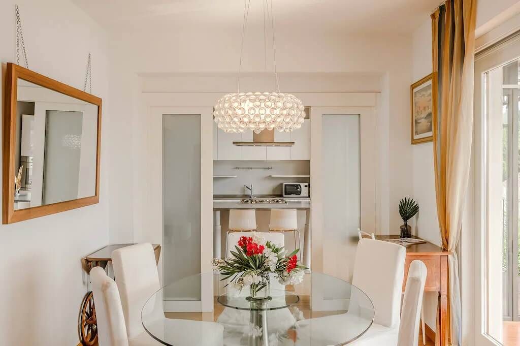 Un coin repas avec une table en verre, des chaises blanches, un lustre et un vase rempli de fleurs.