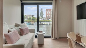 Ein modernes Wohnzimmer mit einer Couch, einem kleinen runden Tisch mit Obst, einem Sessel und einem großen Fenster mit Blick auf Gebäude und Wasser.