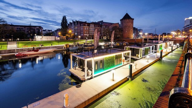 Ein Ufergebiet mit modernen schwimmenden Häusern, einem Kanal und einem historischen Turm im Hintergrund.