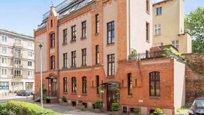 Ein mehrstöckiges Gebäude mit Ziegelaußenwand, einem Balkon im oberen Geschoss und einem Eingangsbereich im Erdgeschoss mit Tür und Fenster.
