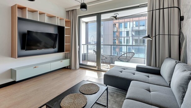 Un soggiorno moderno con un'ampia televisione a schermo piatto, un divano grigio e un balcone con vista su edifici e una ruota panoramica.