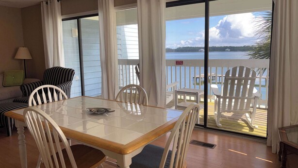 Un coin repas avec une table et des chaises en verre, une vue sur un lac et un balcon avec des sièges d’extérieur.