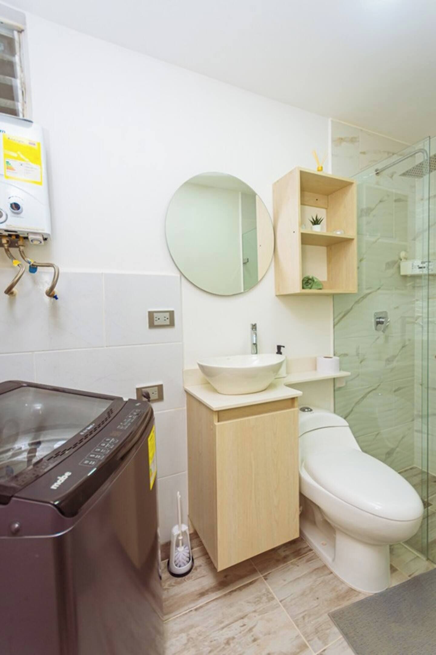 Et badeværelse med vaskemaskine, toilet, håndvask, spejl og brusekabine i glas.