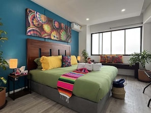 Ein modernes Schlafzimmer mit einem großen Bett, einem farbenfrohen Wandbild, einem Fenster mit Blick und einer Topfpflanze.