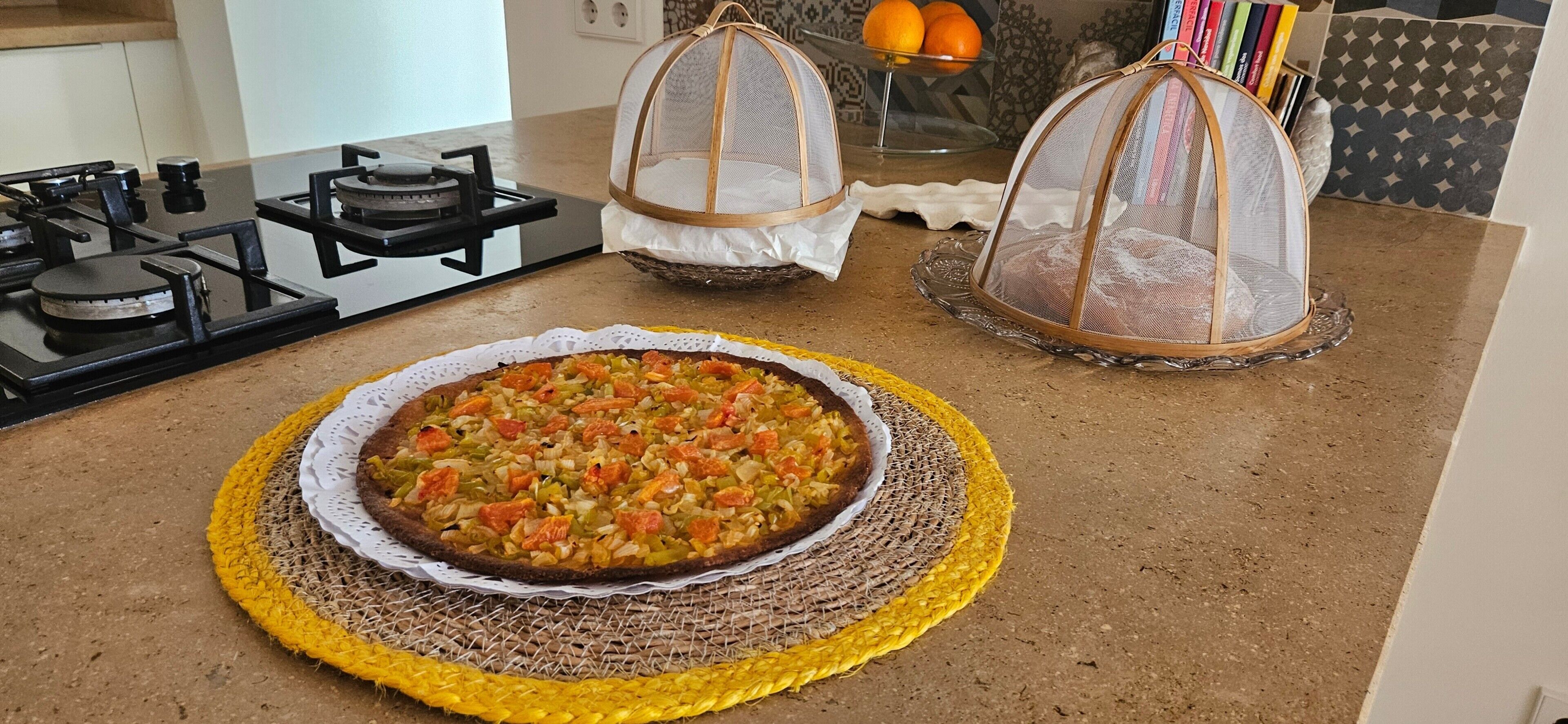 Cozinha com uma pizza sobre um tapete de palha, um fogão a gás e duas cestas de rede suspensas.
