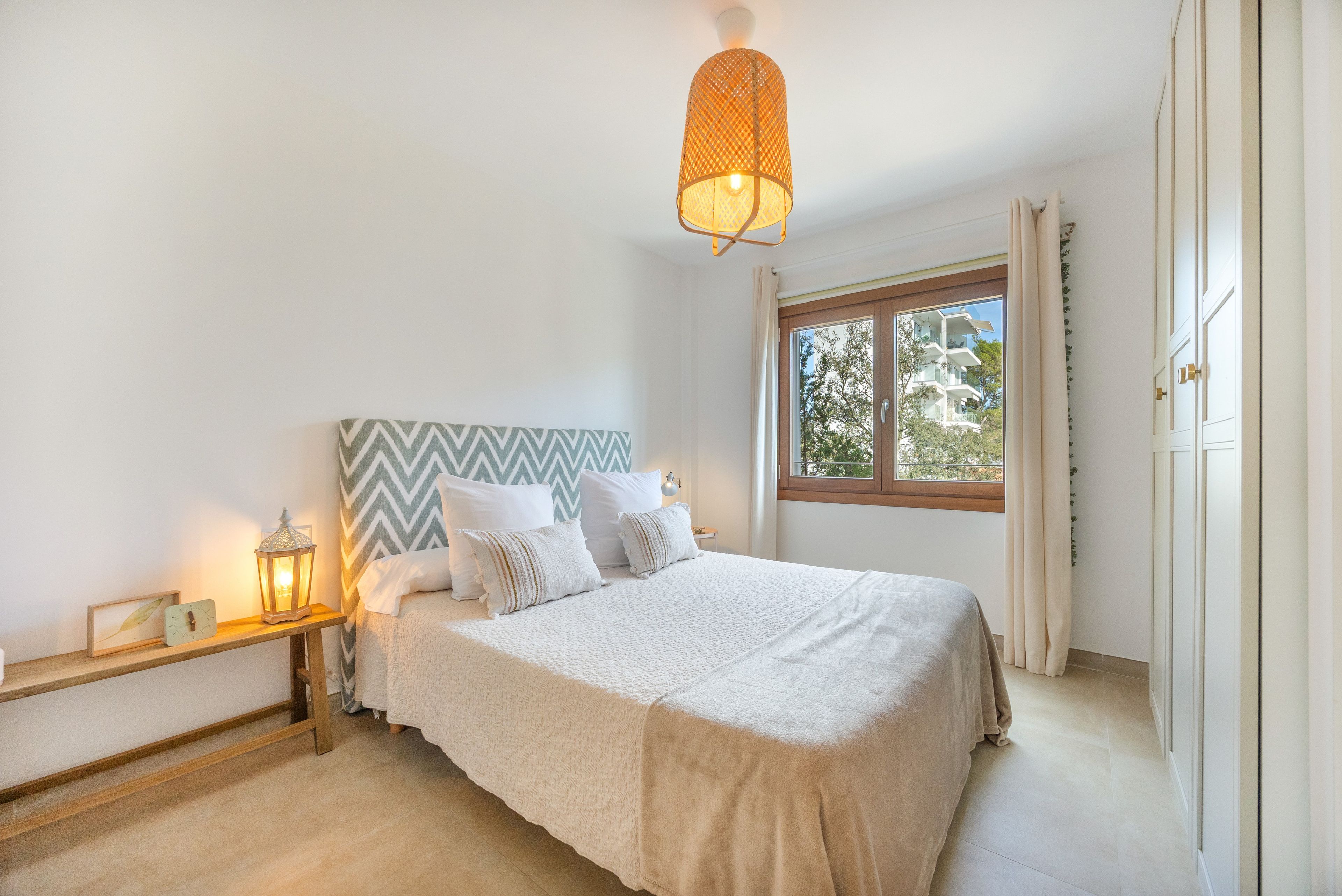 A bedroom with a bed, a nightstand, a wall-mounted lamp, and a window with curtains.