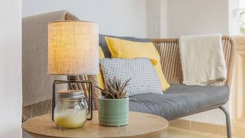 Un rincón acogedor con sillones, una lámpara, una mesita con un frasco y una planta, y un cojín con almohada amarilla.