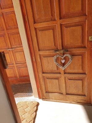 Una puerta de madera con un adorno en forma de corazón, un felpudo texturizado en la entrada y una vista de una habitación con paneles de madera.