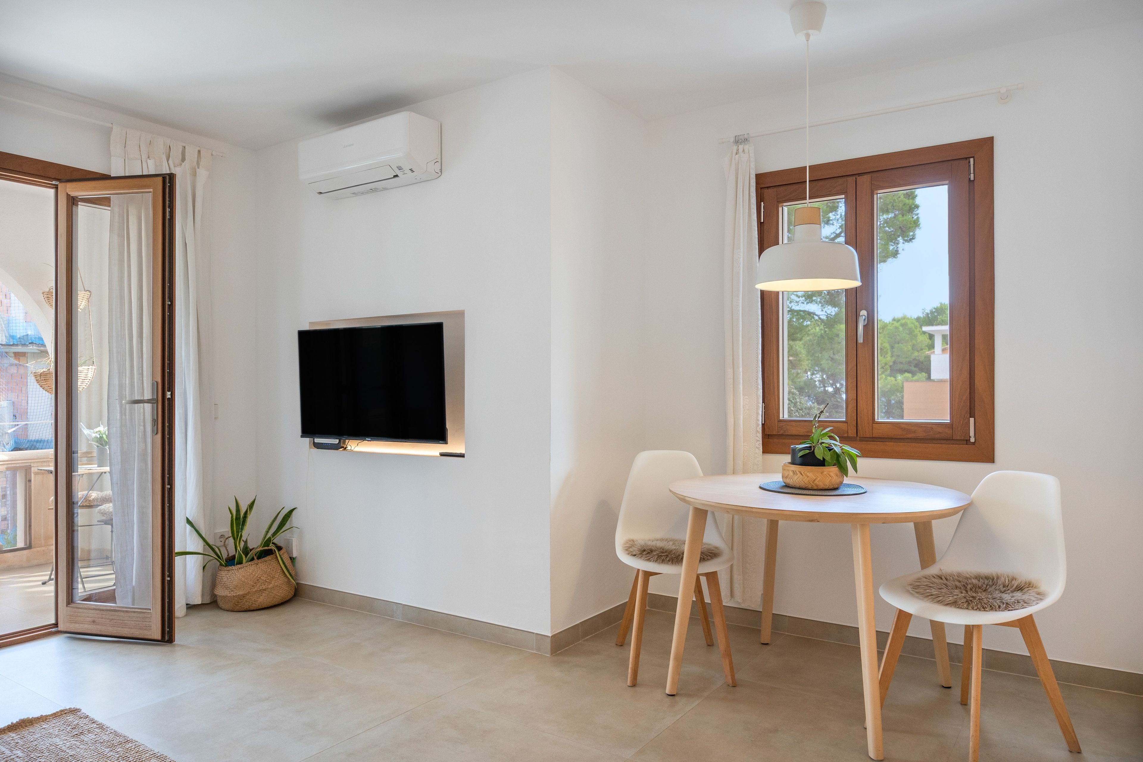 Un salon moderne avec un téléviseur à écran plat fixé au mur, une table à manger avec des chaises, un luminaire suspendu et une fenêtre avec des rideaux.