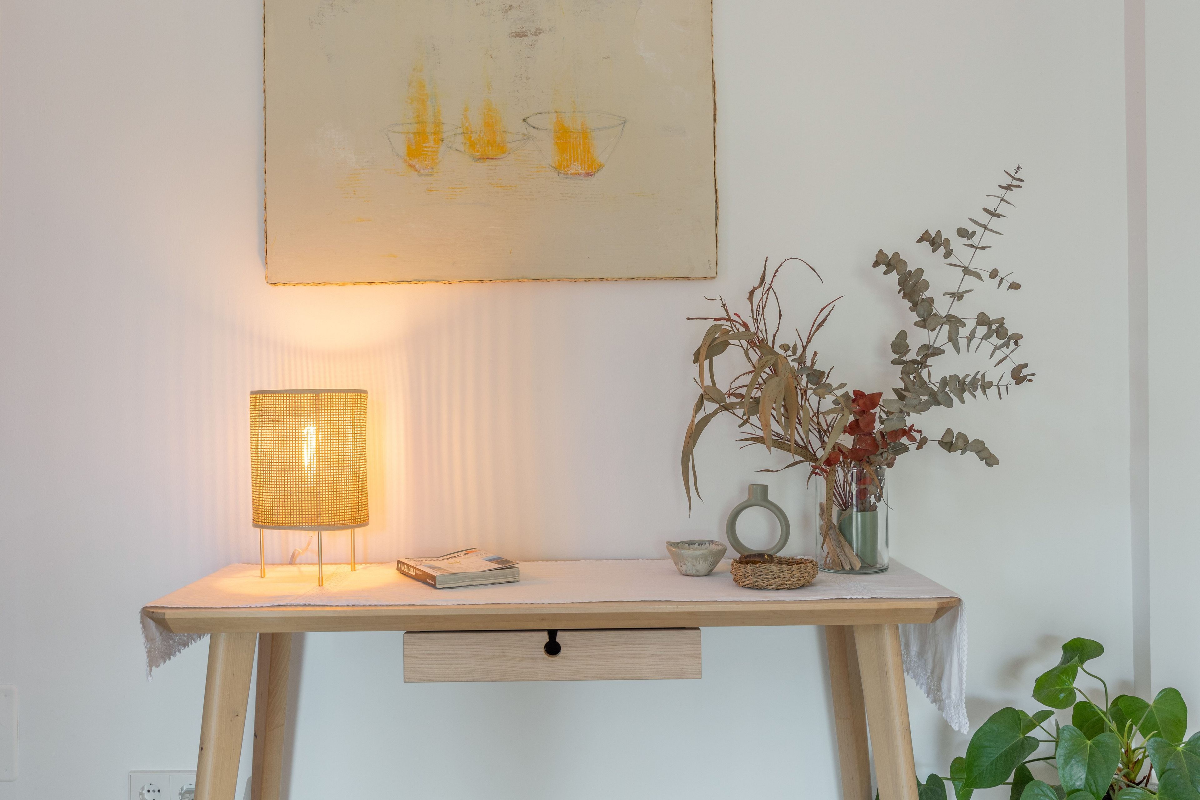 Un bureau en bois, une lampe, un vase contenant des plantes séchées et une œuvre d’art encadrée.