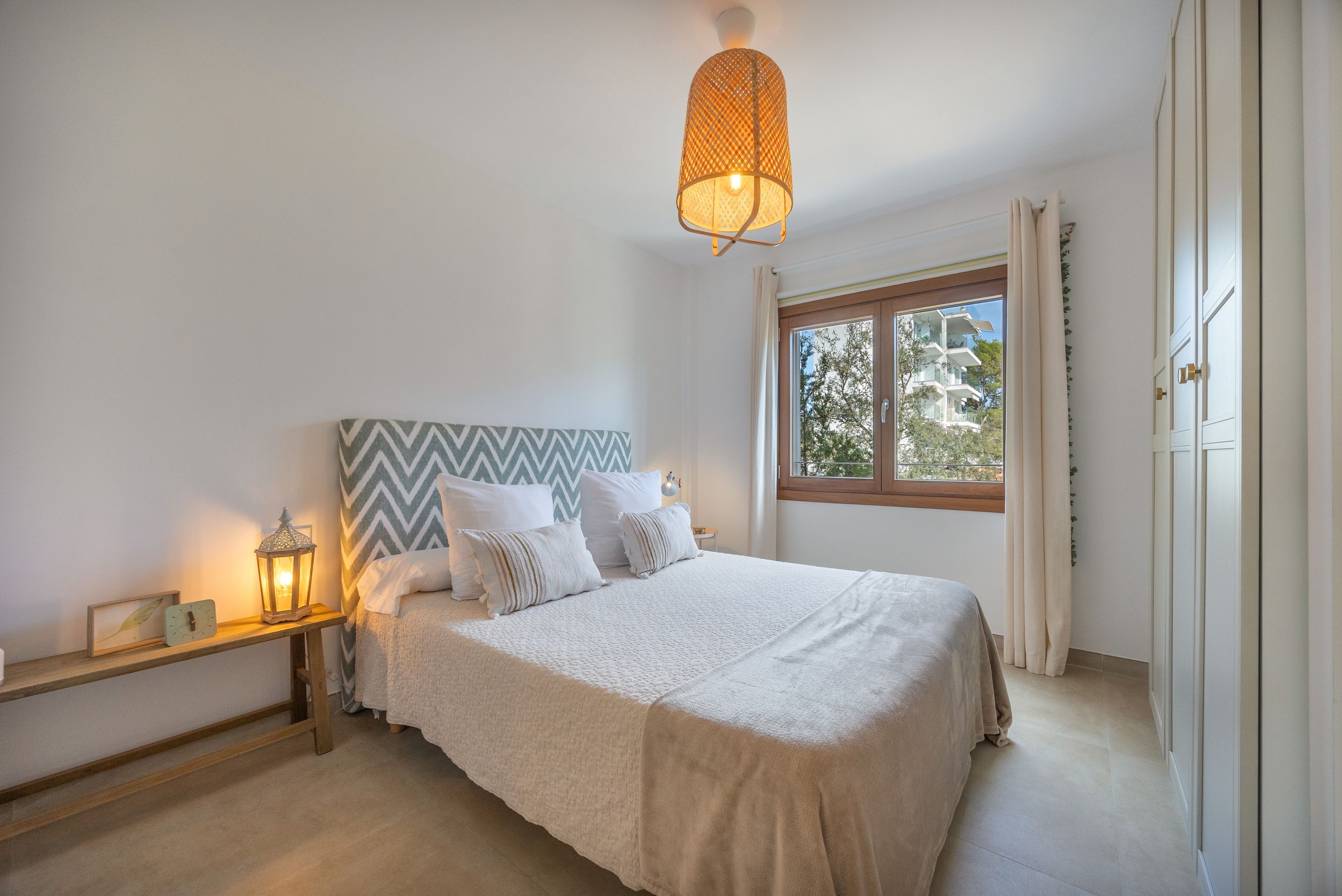Un dormitorio con una cama, una mesita de noche, un reloj de pared, una ventana y una lámpara colgante.