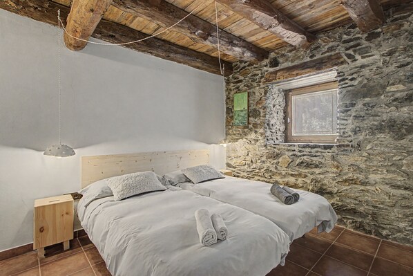 A bedroom with a stone wall, a wooden ceiling, a bed with white bedding, and a window with a view.