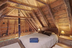An attic bedroom with a wooden ceiling, a bed with pillows, a nightstand with a lamp, and a hanging light bulb.