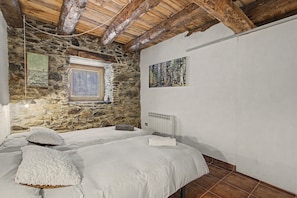 Un dormitorio con pared de piedra, vigas de madera, cama con ropa blanca, una ventana y un cuadro en la pared.