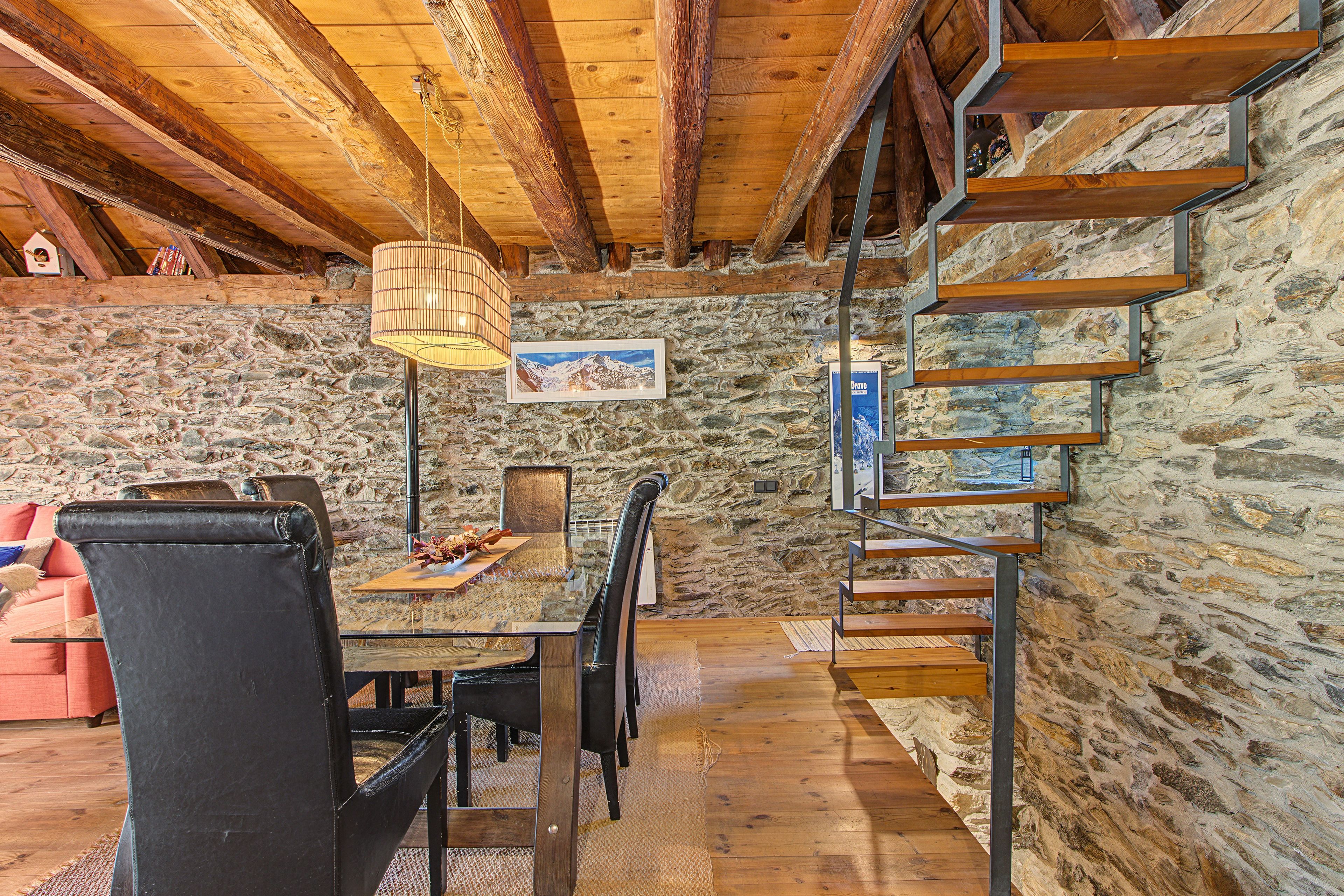 Un interior rústico con pared de piedra, escalera de madera y un comedor con mesa y sillas.