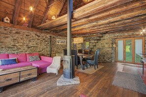 A rustic interior with a stone wall, wooden beams, a pink sofa, a fireplace, and a dining area.