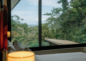 Une chambre dotée d’une grande fenêtre donnant sur une forêt luxuriante et offrant une vue lointaine sur l’océan.