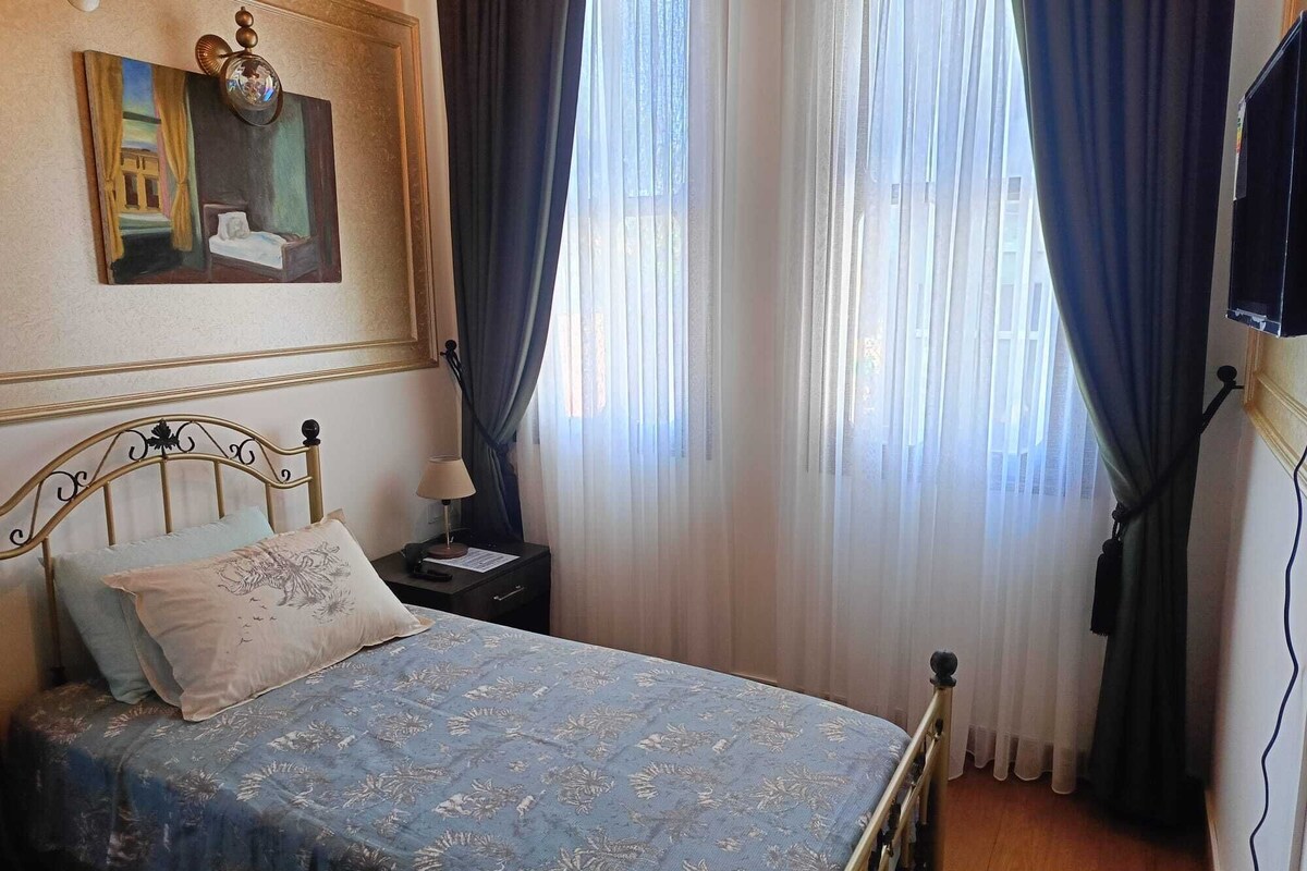 A bedroom with a bed, a nightstand, a lamp, a painting on the wall, and a window with curtains.