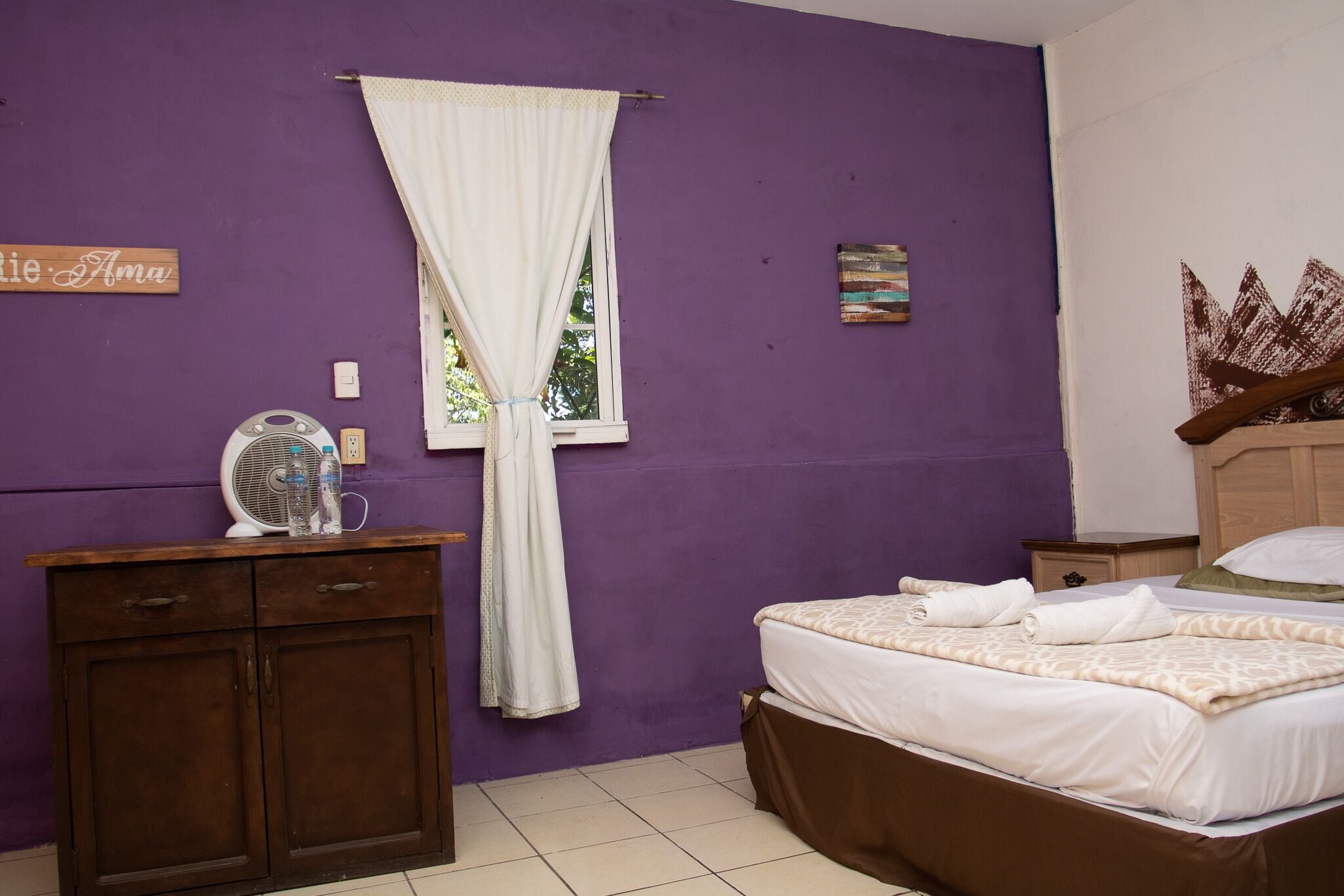 Quarto com cama, cômoda, ventoinha e janela com cortinas.