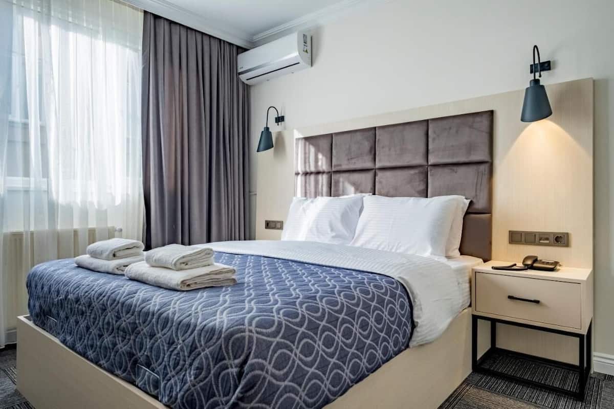 A hotel room with a bed, two bedside lamps, a nightstand with a phone, and a window with curtains.