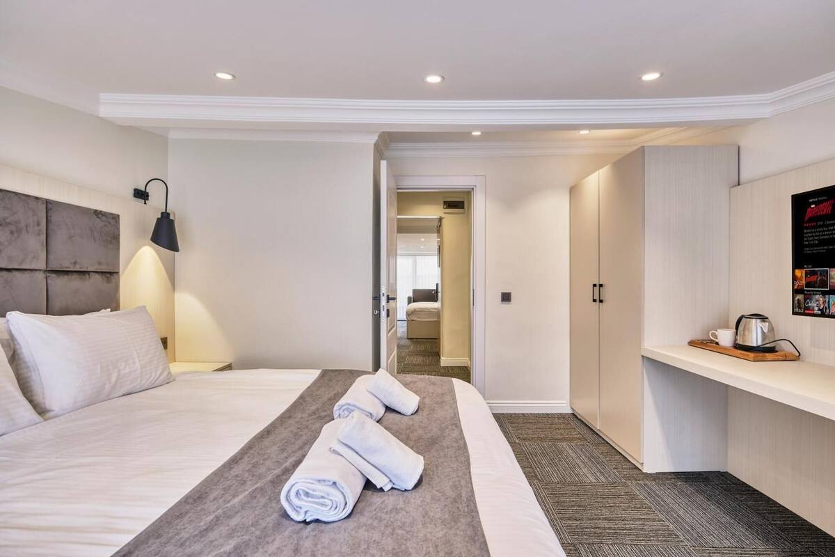A hotel room with a bed, a bedside lamp, a towel rack, a small table with a cup, and a wall-mounted TV.