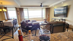 A hotel room with a dining area, a living room with a sofa and a TV, and a view of the outdoors. - Calafia Resorts and Villas | Oceanfront Condo, Pools, Hot Tubs & Wellness Room 🌊 (Playas de Rosarito)