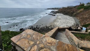 A coastal view with a stone staircase leading to a beach, a rocky shoreline, and distant buildings. - Calafia Resorts and Villas | Oceanfront Condo, Pools, Hot Tubs & Wellness Room 🌊 (Playas de Rosarito)