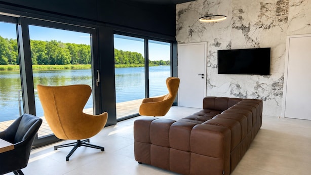 A modern living room with a sofa, two armchairs, a television, and a view of a lake through large windows. - Dziwnow Boat House With Jacuzzi (Dziwnów)