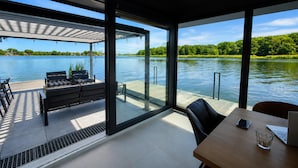 A modern outdoor deck with a view of a lake, furnished with a table and chairs, and a covered seating area. - Dziwnow Boat House With Jacuzzi (Dziwnów)