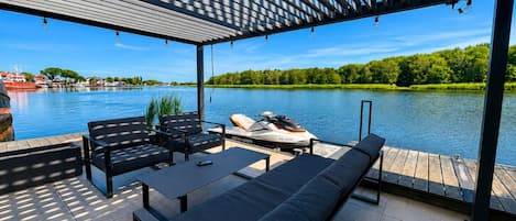 Un espace extérieur abrité, aménagé avec des sièges, et offrant une vue sur un lac et des bateaux.