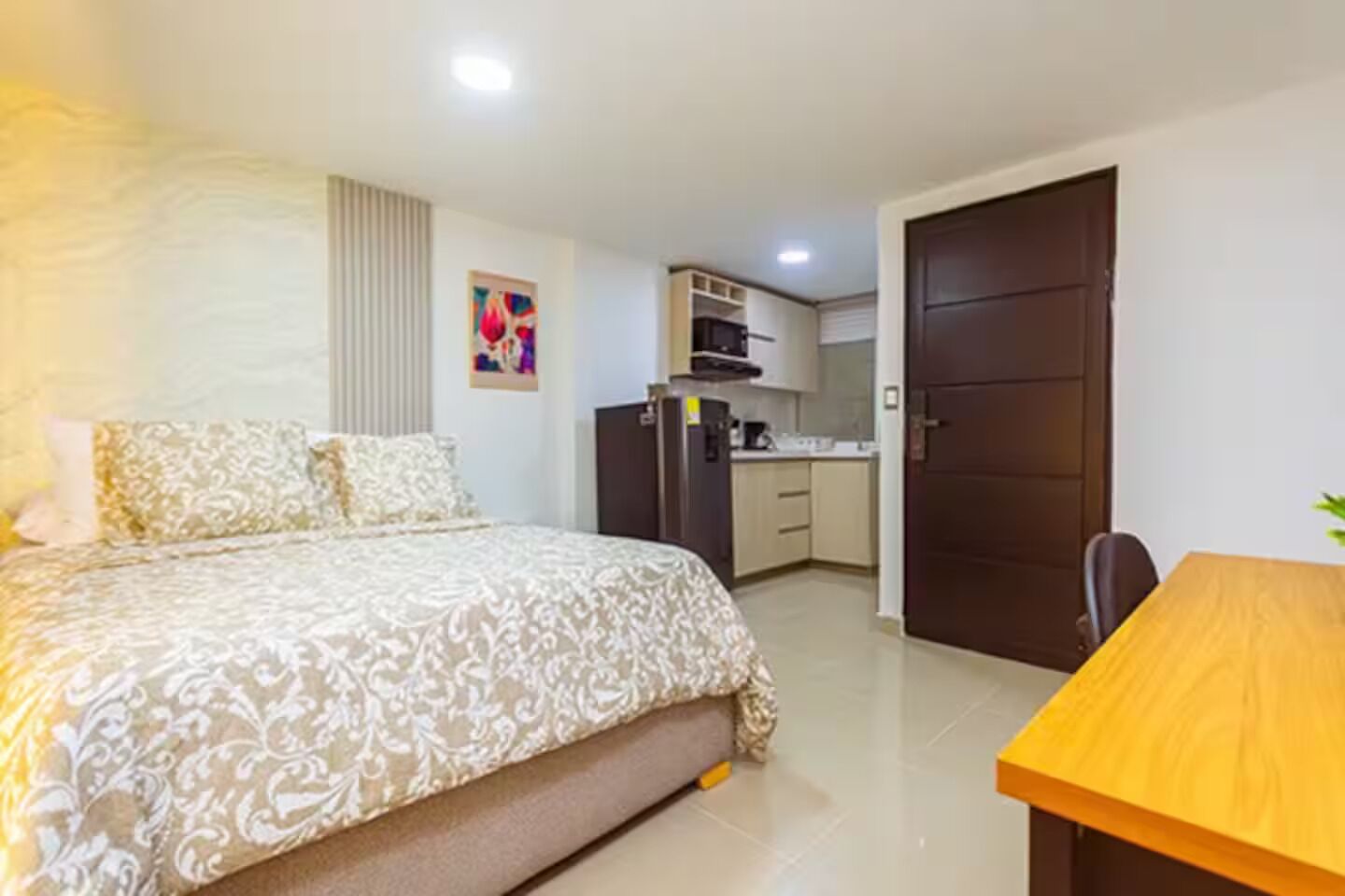 A bedroom with a bed, a desk, a kitchenette, and a door.