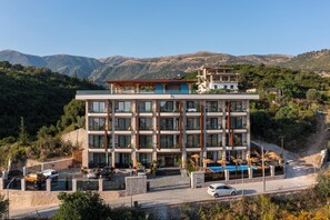 A multi-story hotel with a modern design, featuring balconies, a pool area, and a view of the surrounding hills.