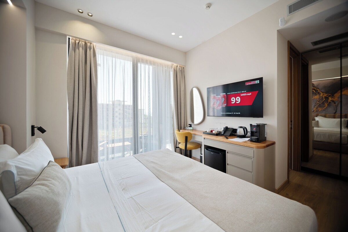 Una habitación de hotel con una cama, un escritorio con cafetera, un televisor y una ventana con cortinas.