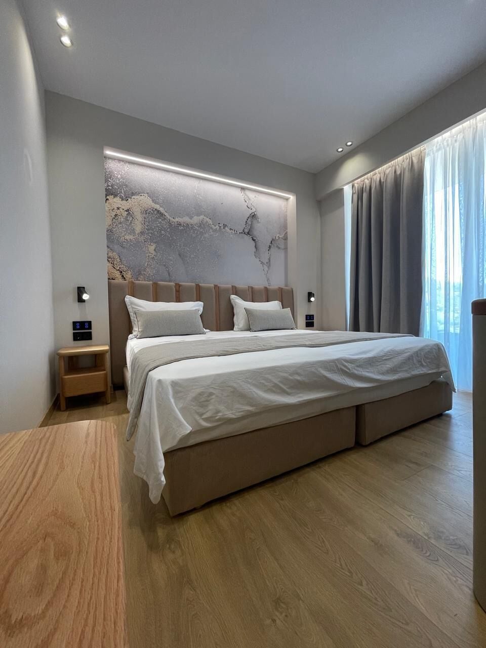 A modern bedroom with a large bed, a nightstand, a wall-mounted light, and a window with curtains.