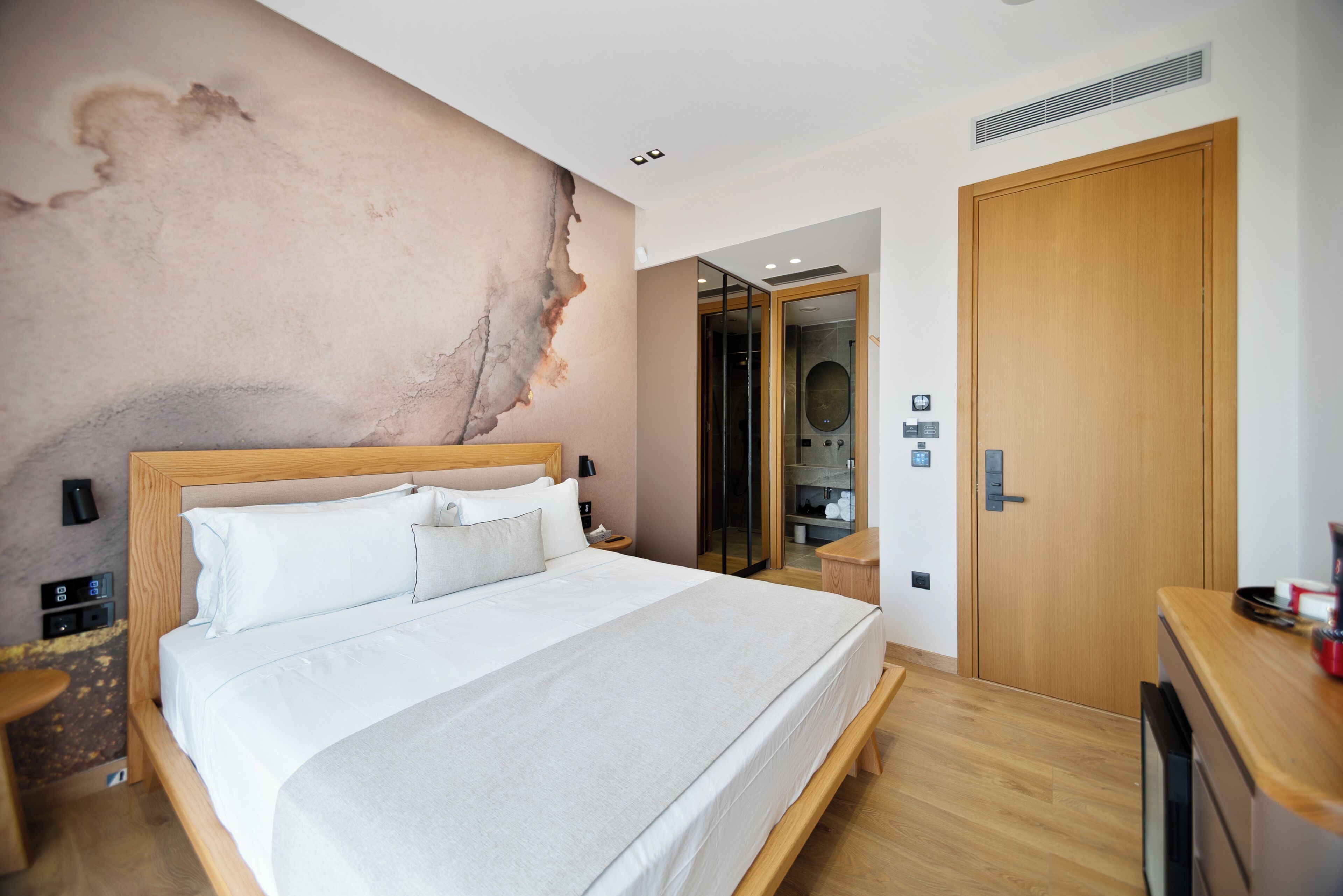 A modern hotel room with a large bed, a wooden headboard, a built-in wardrobe, a bathroom with a glass shower, and a small desk with a chair.